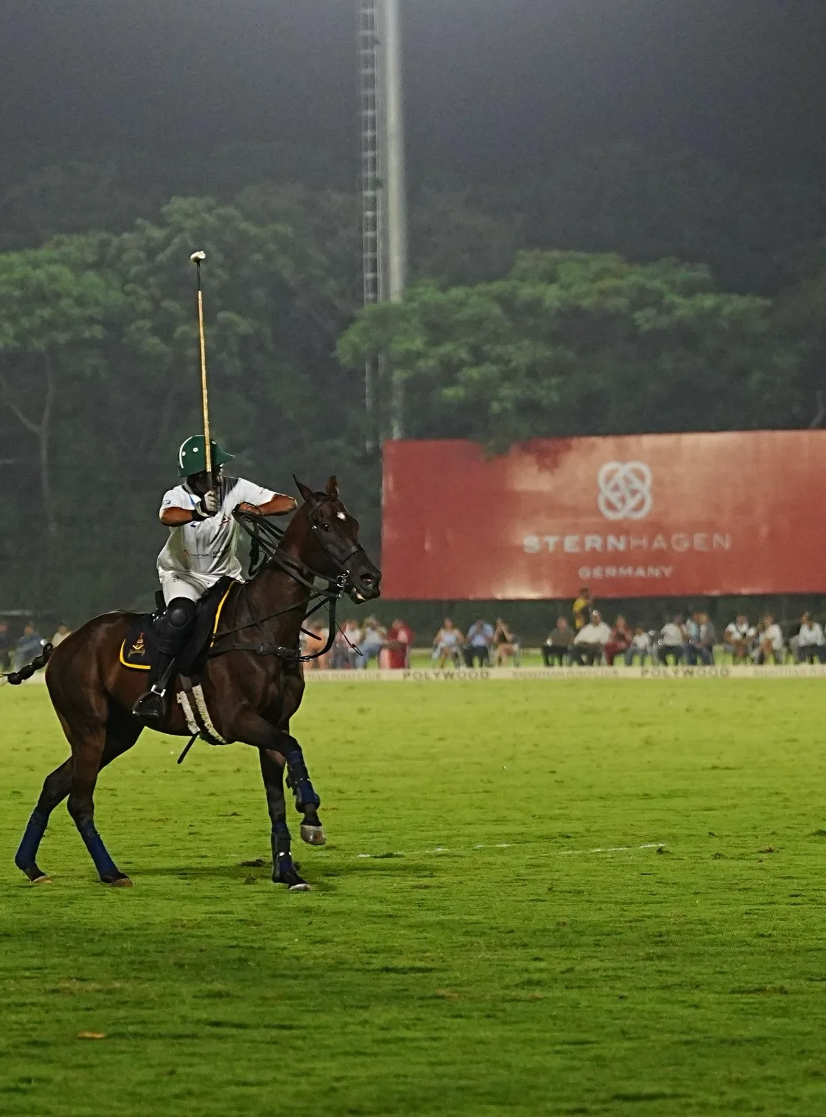 A polo player in a green helmet raises a mallet on horseback during a floodlit evening match, in front of a Sternhagen Germany billboard